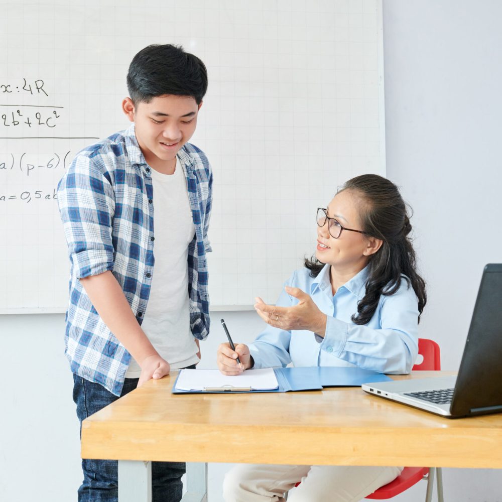 Smiling mature math teacher explaining difficult topic to confused schoolboy after class